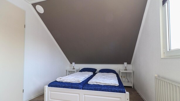 Ein kleines Zimmer mit einem Einzelbett, geneigter Decke, weißen Wänden und einem Fenster.