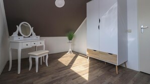 Ein Zimmer mit weißem Schminktisch, einem Spiegel, einem kleinen Hocker, einer Topfpflanze und einem Kleiderschrank mit Holdbase.