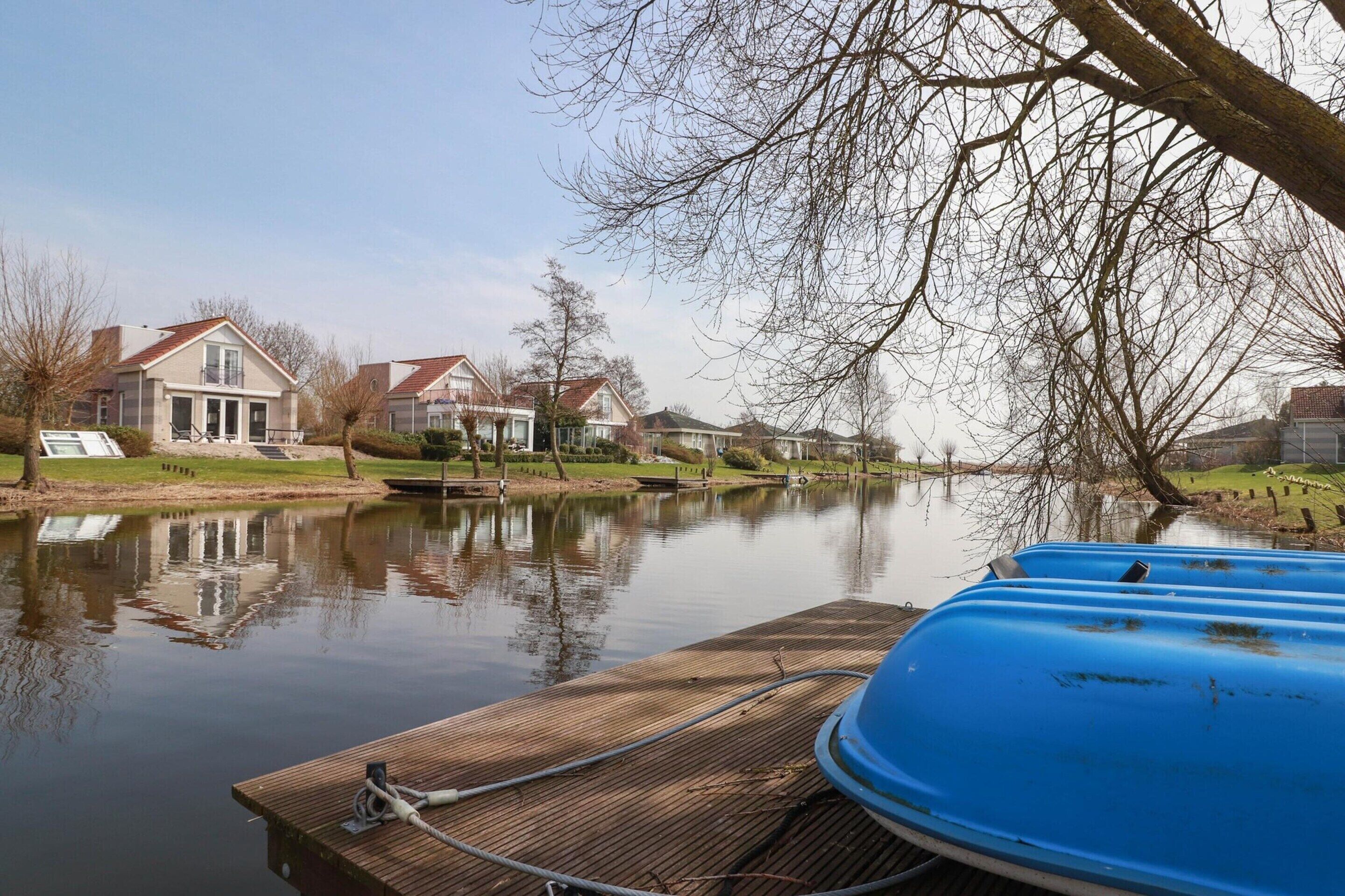A calm river with houses on the opposite bank, a dock with a boat, and a clear sky.