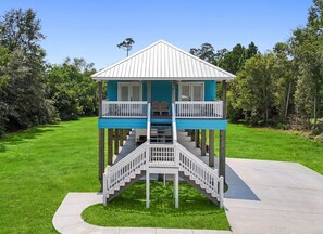 A two-story wooden structure with a balcony on each level, a staircase connecting the floors, and a surrounding grassy area. - Beautiful 4-bedroom house with AC in quaint Waveland (Waveland)