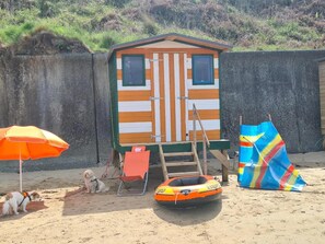 Ein Strandhaus mit einer kleinen Terrasse, einem Liegestuhl, einem Hund und einem farbenfrohen Strandschirm.