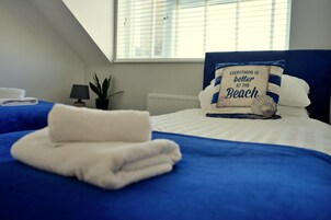 Habitación de hotel con cama, almohada con motivos playeros, toalla doblada, lámpara y planta en maceta.