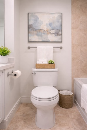 A bathroom with a toilet, a towel rack with towels, a framed abstract painting, a potted plant, and a wicker basket. - Spacious 2BR Apartment in Central Toronto (Toronto)