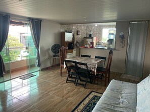 Ein Essbereich mit Tisch und Stühlen, ein Ventilator und eine Küchenzeile mit Kühlschrank, Herd und Kaffeemaschine.