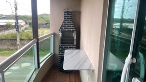 A balcony with a brick barbecue grill, a white marble countertop, and a glass door leading to a view of a street and greenery.