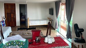 A room with a bed, a table with a kettle, a fan, and a window with curtains.