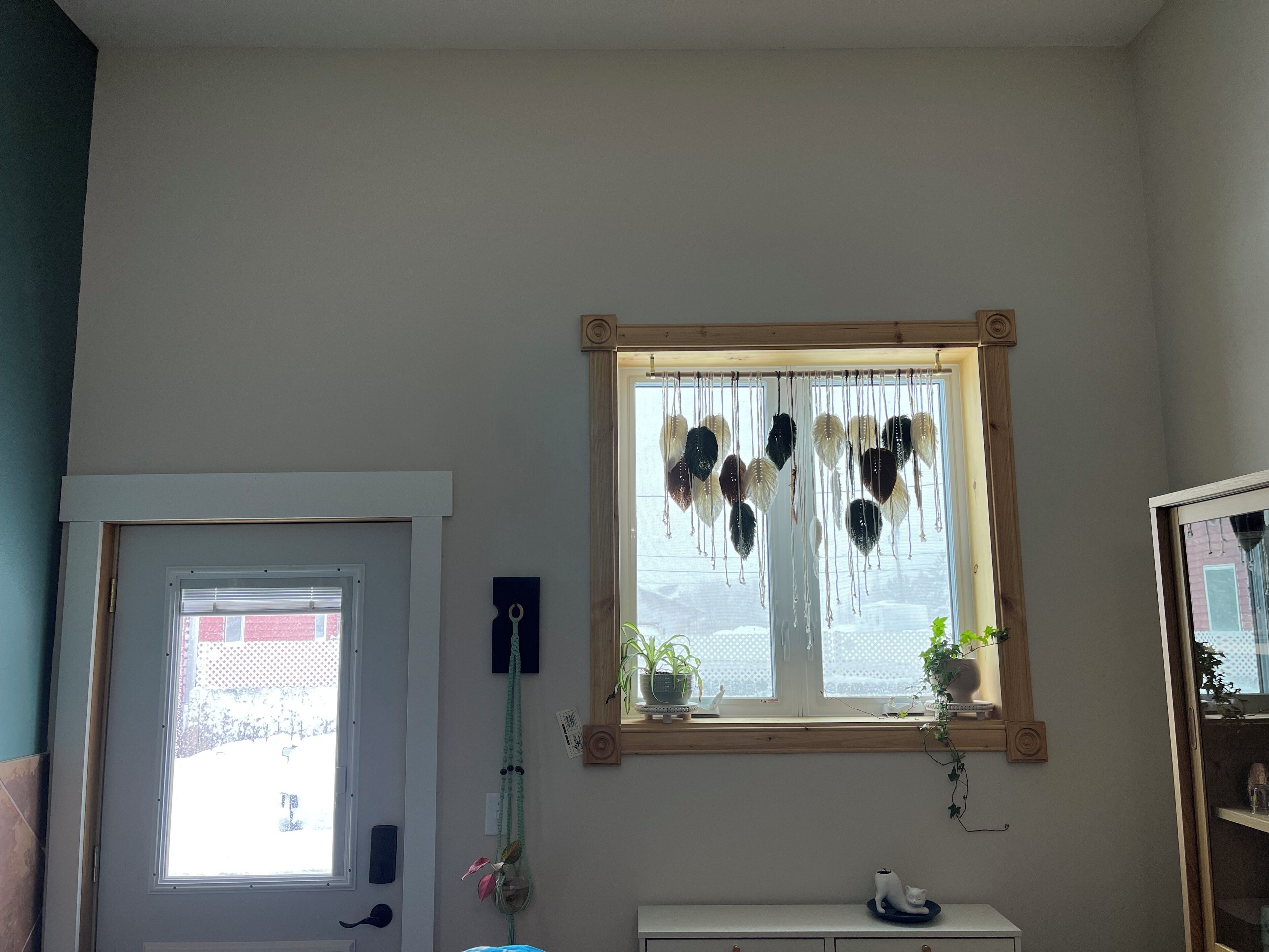 A room with a window, a door, a small shelf with a plant, and a wall-mounted decoration.