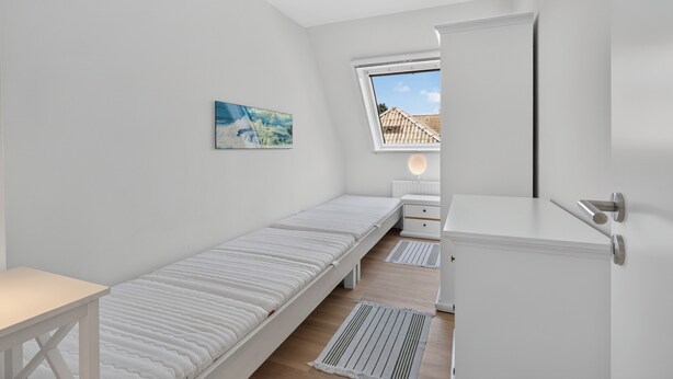 Una habitación pequeña con cama individual, blanca, con ventana, mesita de noche, lámpara y armario.