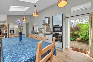 Uma cozinha moderna com eletrodomésticos de aço inoxidável, uma área de jantar com toalha de mesa estampada e vista para um jardim através de uma porta deslizante.