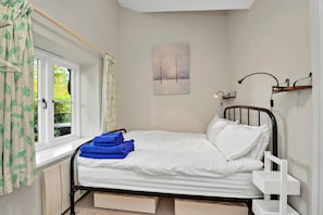 Uma cama de solteiro com roupa de cama branca e toalhas azuis, uma janela com cortinas, um quadro na parede e uma mesa de cabeceira com um abajur.