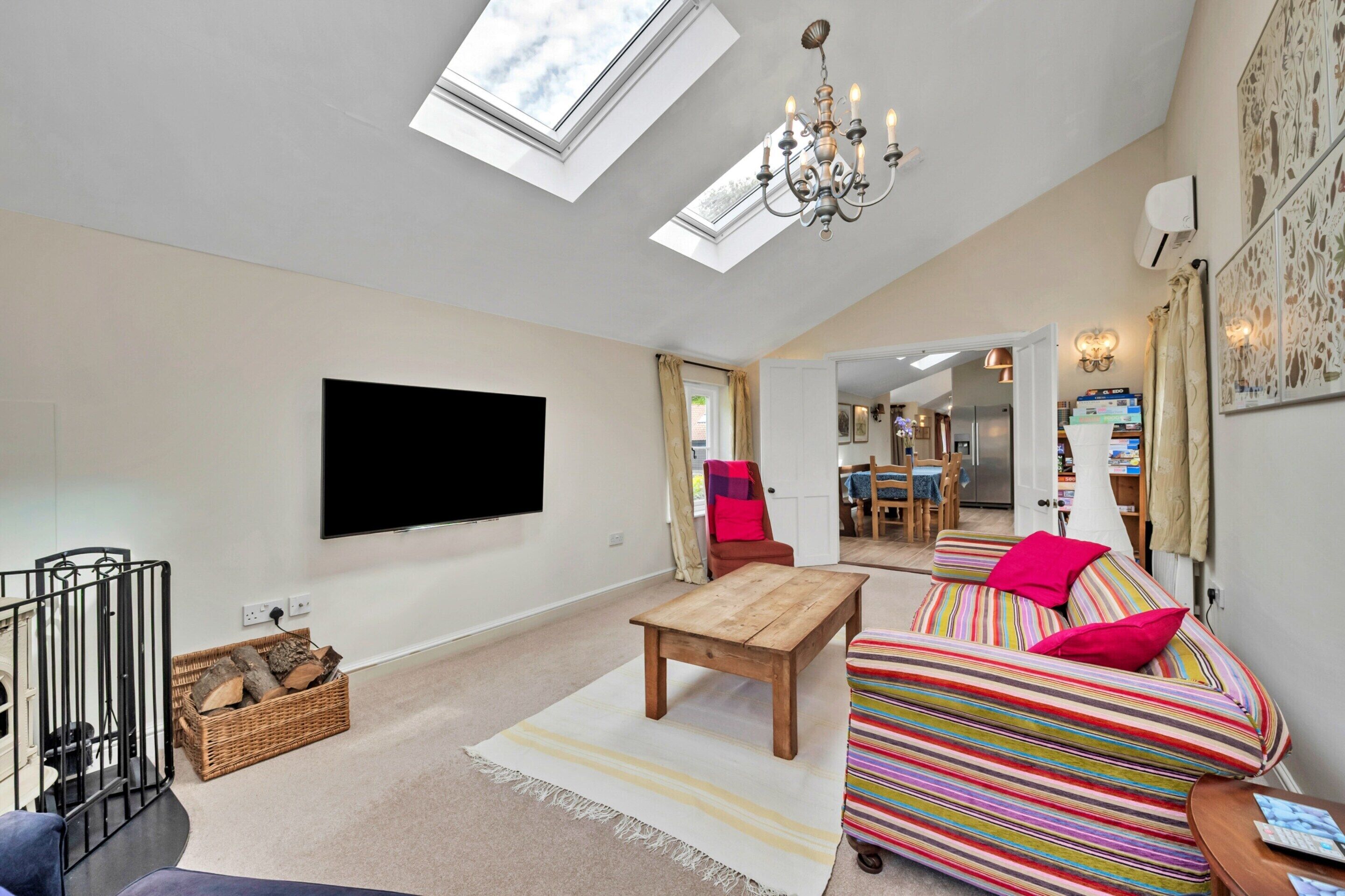 A modern living room with a flat-screen TV, a wooden coffee table, a striped sofa, a chandelier, and a skylight.