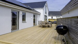 A covered outdoor patio with a barbecue grill, wooden deck, and a view of neighboring houses. - Stylish Barn Retreat for 8 (Pwllheli)