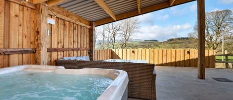 A wooden spa area with a hot tub and wicker seating, overlooking a rural landscape.
