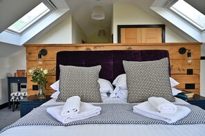 Une chambre avec un plafond en pente, une tête de lit en bois, deux grands coussins à motifs, une table de chevet avec une lampe et un vase rempli de fleurs.