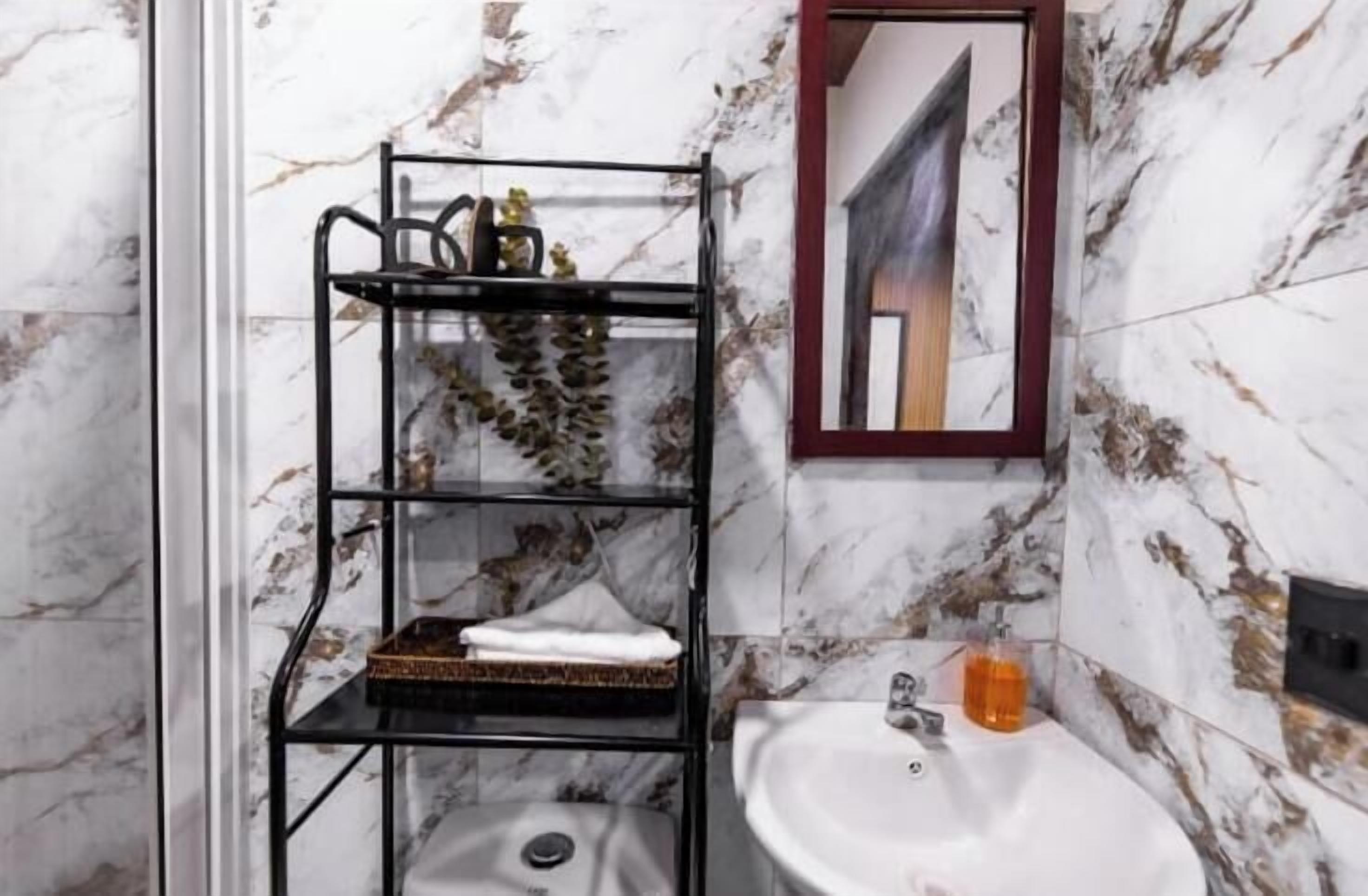 Un baño con pared de mármol, un estante de metal negro con toallas, un lavabo y un espejo.