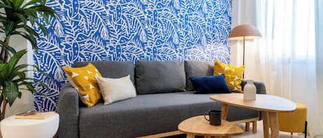 En moderne stue med en grå sofa, gule og blå puder, et lille træbord og en gulvlampe.
