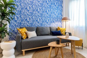 Een moderne woonkamer met een grijze bank, gele en blauwe kussens, een klein houten tafeltje en een vloerlamp.