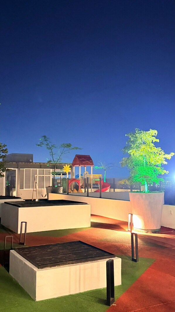 Un terrain de jeux équipé d’un toboggan et de structures d’escalade, entouré d’un espace paysager agrémenté d’un grand arbre et d’un bâtiment à toit rouge.