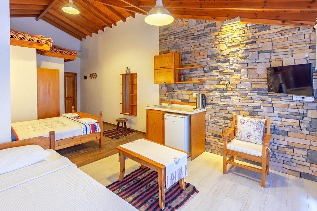 A cozy room with a bed, a wooden bench, a chair, a small table, a kitchenette, and a stone wall.
