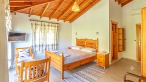 A bedroom with a wooden ceiling, a bed with a wooden headboard, a bedside table, a chair, a television, and a window with curtains.
