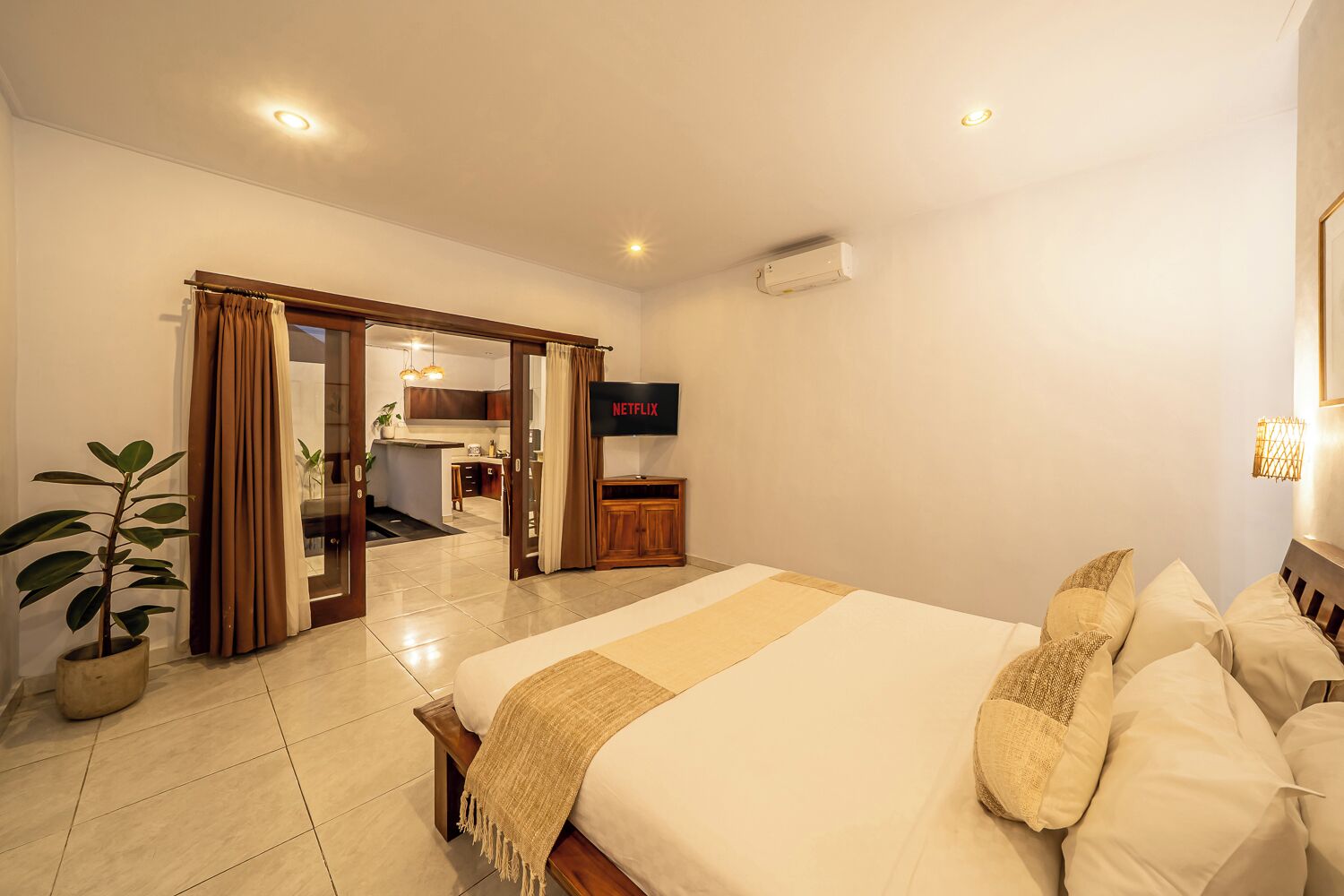 A hotel room with a bed, a television, a potted plant, and a view into the kitchen area.