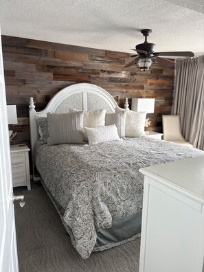 A bedroom with a wooden wall panel, a white headboard, a patterned bedspread, and a ceiling fan. - Luxurious 4-Bedroom 4-Bathroom Condo North Myrtle Beach Oceanfront (North Myrtle Beach)