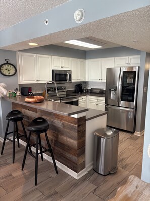A modern kitchen with white cabinets, stainless steel appliances, a wooden island with bar stools, and a trash can. - Luxurious 4-Bedroom 4-Bathroom Condo North Myrtle Beach Oceanfront (North Myrtle Beach)