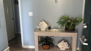 Un pasillo con una consola de mimbre, una lámpara de pared, una planta en maceta y una puerta.