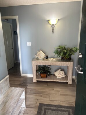 A hallway with a wicker console table, a wall-mounted light fixture, a potted plant, and a door. - Luxurious 4-Bedroom 4-Bathroom Condo North Myrtle Beach Oceanfront (North Myrtle Beach)