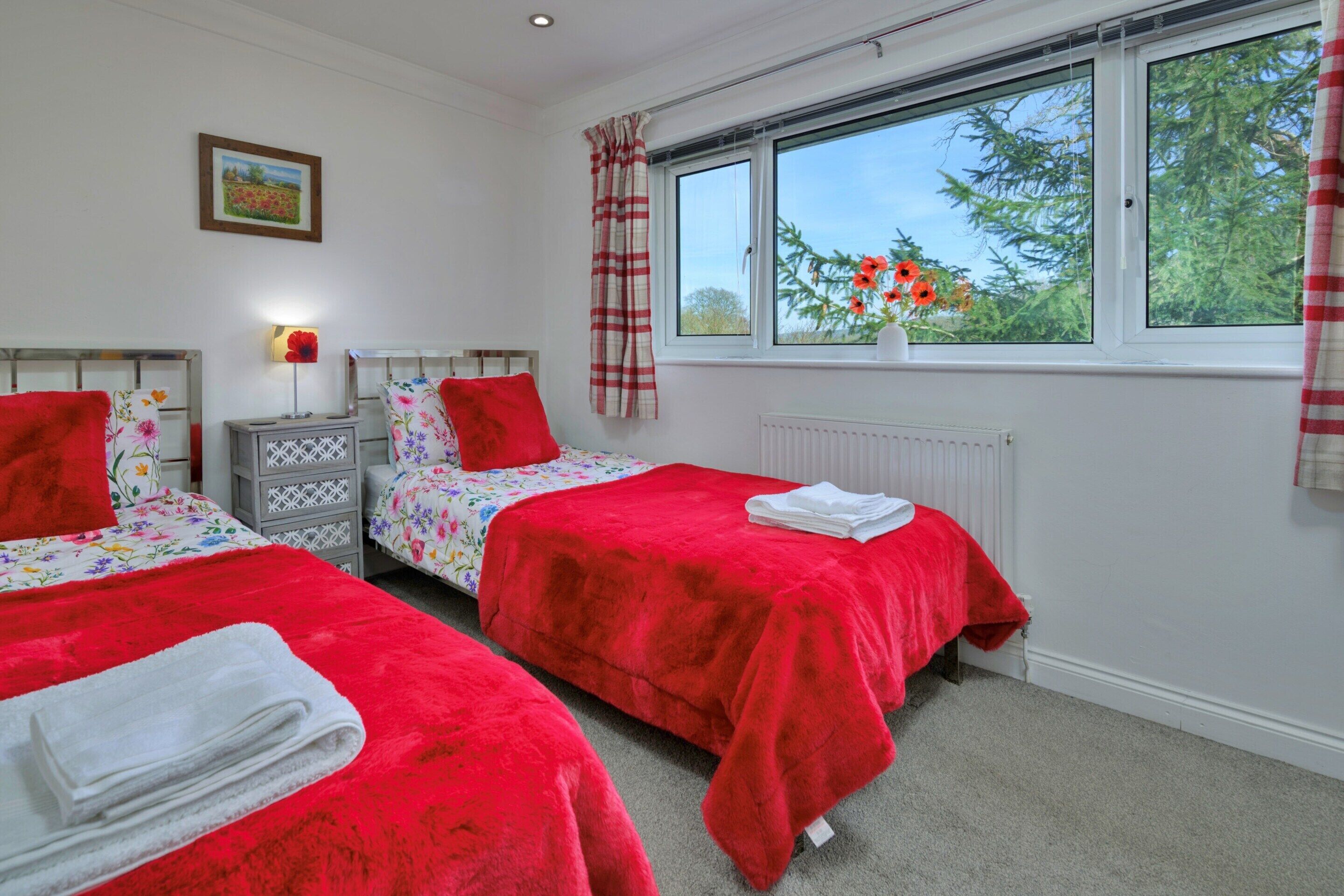 A bedroom with two beds, a nightstand, a lamp, a window with a view of trees, and a radiator.