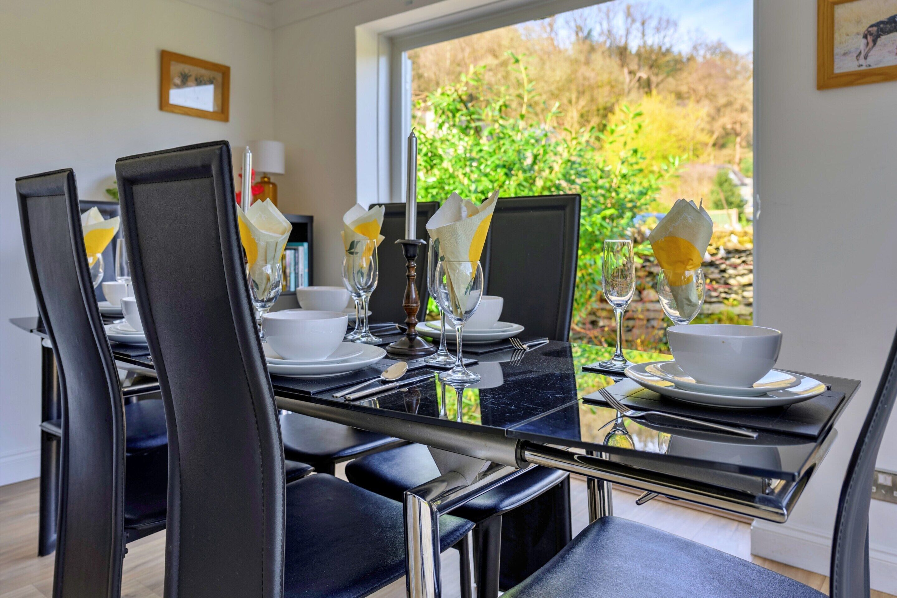 A dining table set with chairs, plates, cutlery, and glasses, with a view of greenery outside.