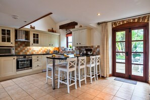 A kitchen with a central island, white bar stools, and a large window with a view of greenery. - Luxury Whitby Townhouse for 10 (Bude)