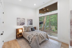 A bedroom with a large window, a bed with a grey duvet, two wall-mounted paintings, and a bedside table with a lamp. - Luxury Mountain Getaway Minutes from Crystal Mountain & Mount Rainier (Enumclaw)