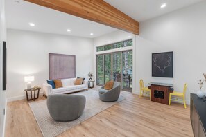 Ein modernes Wohnzimmer mit einem weißen Sofa, zwei grauen Sesseln, einer Holzbalkendecke und einem großen Fenster mit Blick auf Bäume.