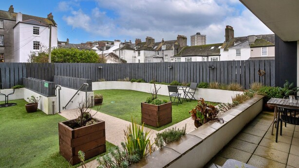 Patio trasero residencial con césped, jardineras de madera, un espacio de descanso con sillas y vista a casas vecinas.