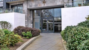 Entrada de un edificio moderno con fachada de piedra, puertas de cristal y un pequeño jardín con arbustos.