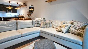 Una sala de estar moderna con un sofá modular, una cocina al fondo y una estantería de madera con objetos decorativos.