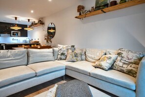 Ein modernes Wohnzimmer mit Ecksofa, einer Küche im Hintergrund und einem Holzregal mit Dekoration.