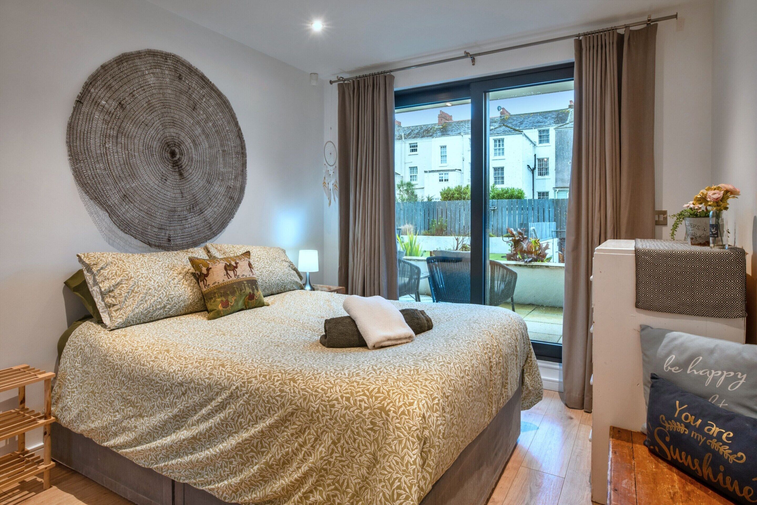 Une chambre à coucher avec un lit, une grande tapisserie murale, une vue sur l’extérieur et une petite table sur laquelle est posé un vase.