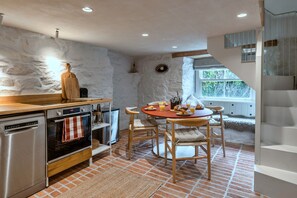 A kitchen with a stone wall, wooden furniture, and a dining area with a view of greenery. - Kingsand Coastal Retreat for 6 (Torpoint)