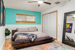 Een slaapkamer met een bed, een plafondventilator, een raam met jaloezieën, een kledingkast en een spiegel.