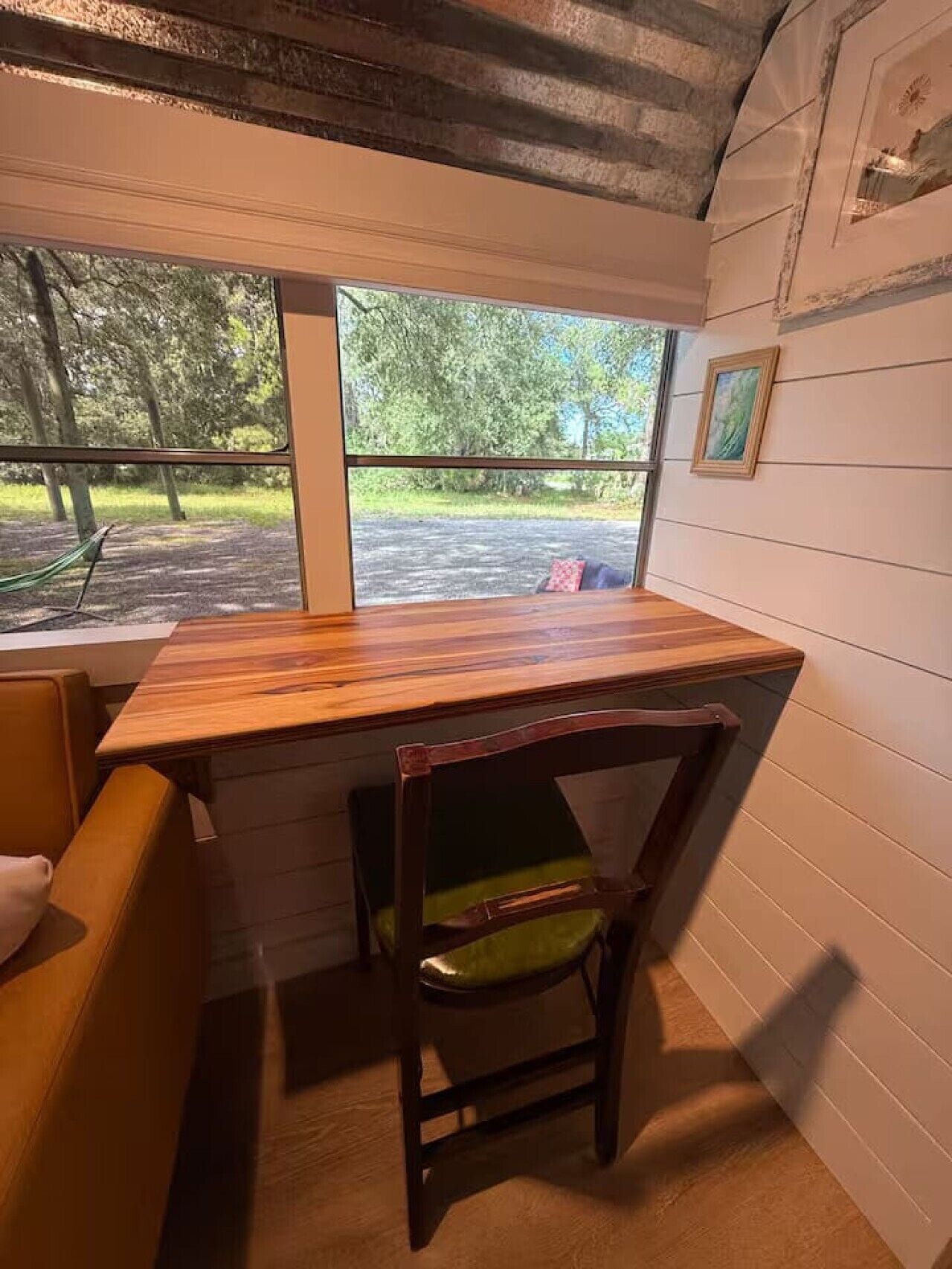 A room with a wooden desk and a chair, a window with a view of a park, and a wall-mounted picture.