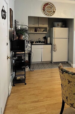 Eine kleine Küche mit weißen Schränken, einem weißen Kühlschrank und einem schwarzen Metallregal mit Gegenständen darauf.