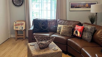 A living room with a brown leather sofa, a woven ottoman, a coffee sign, a wall-mounted clock, a deer head decoration, and a window with curtains.