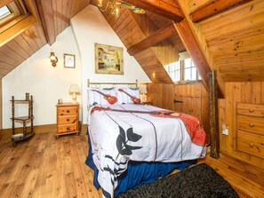 A bedroom with a sloped ceiling, wooden walls, a bed with a floral bedspread, a nightstand, a lamp, and a framed picture on the wall. - 2-bedroom Seaside Home (Whitby)