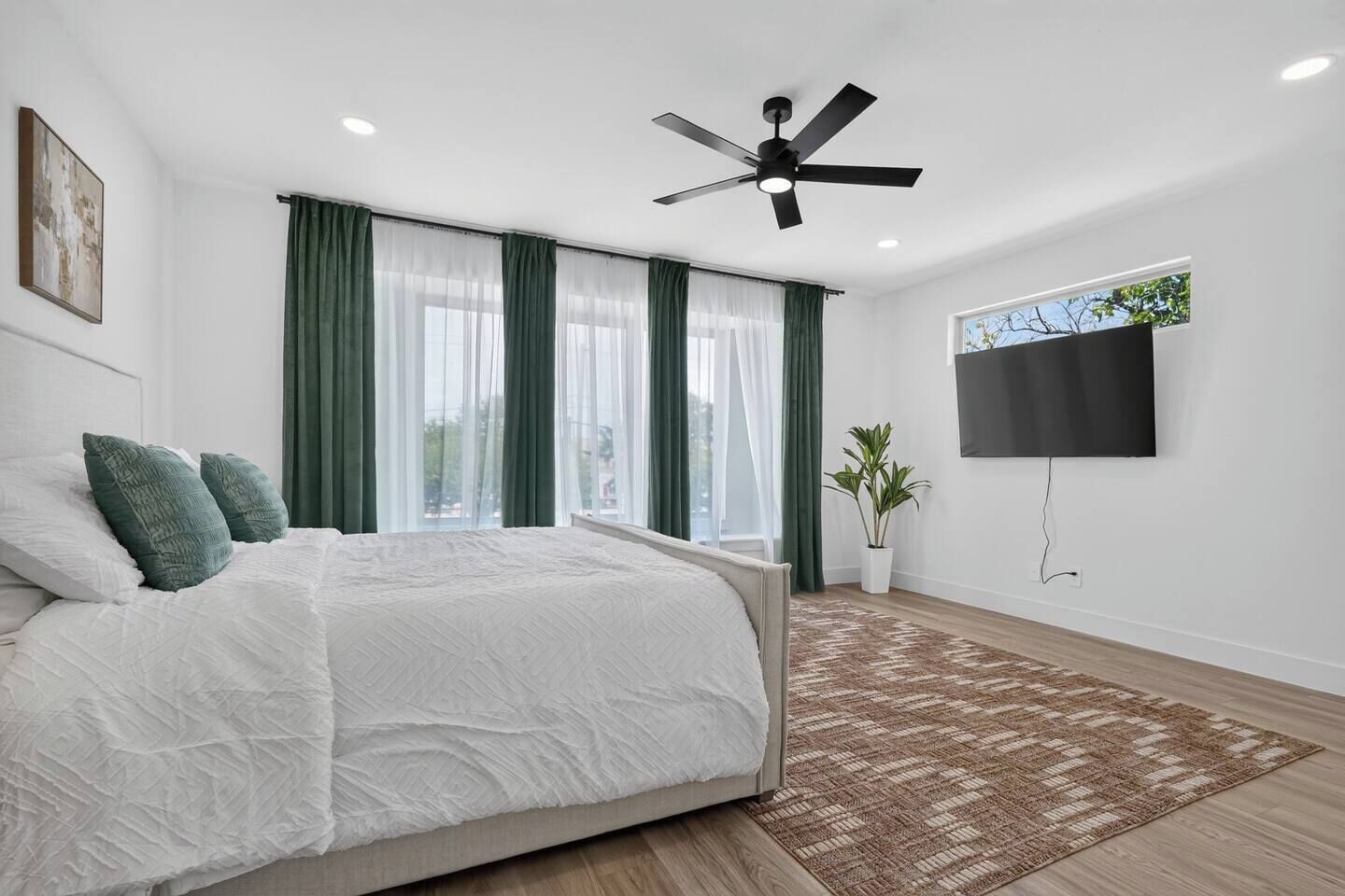 A modern bedroom with a bed, green curtains, a ceiling fan, a TV, and a plant.