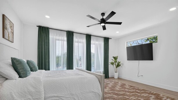 Quarto moderno com cama, cortinas verdes, ventilador de teto, TV e uma planta.