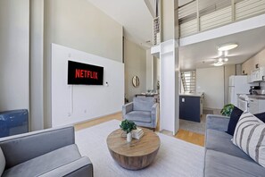 A modern living room with a mounted TV displaying 'NETFLIX,' a dining area with a round wooden coffee table, and a kitchen with white appliances. - Explore Gaslamp from Chic Loft +Balcony City Views (San Diego)