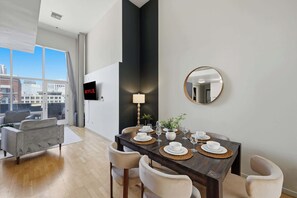 A modern dining area with a dark wooden table set for four, surrounded by cushioned chairs. A television is mounted on the wall, and a round mirror hangs above the table. - Explore Gaslamp from Chic Loft +Balcony City Views (San Diego)