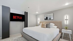 A modern bedroom with a large bed, a wall-mounted TV displaying the Netflix logo, a bedside table with a lamp, and a mirror. - Explore Gaslamp from Chic Loft +Balcony City Views (San Diego)